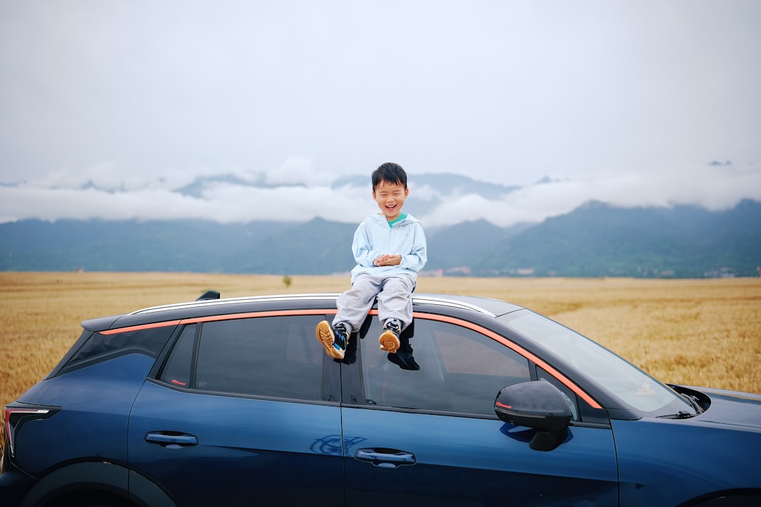 A boy sits on a blue car in a field.